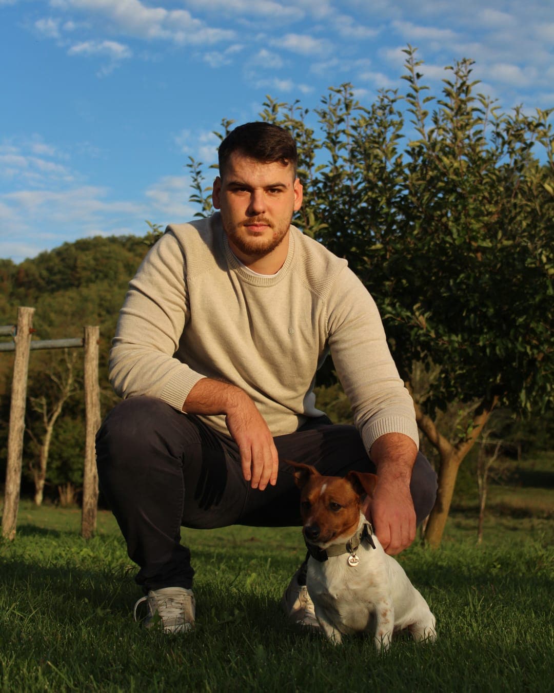 Matej Ritoša čučne na travi uz svog psa, jack russell terijera, ispred voćke.